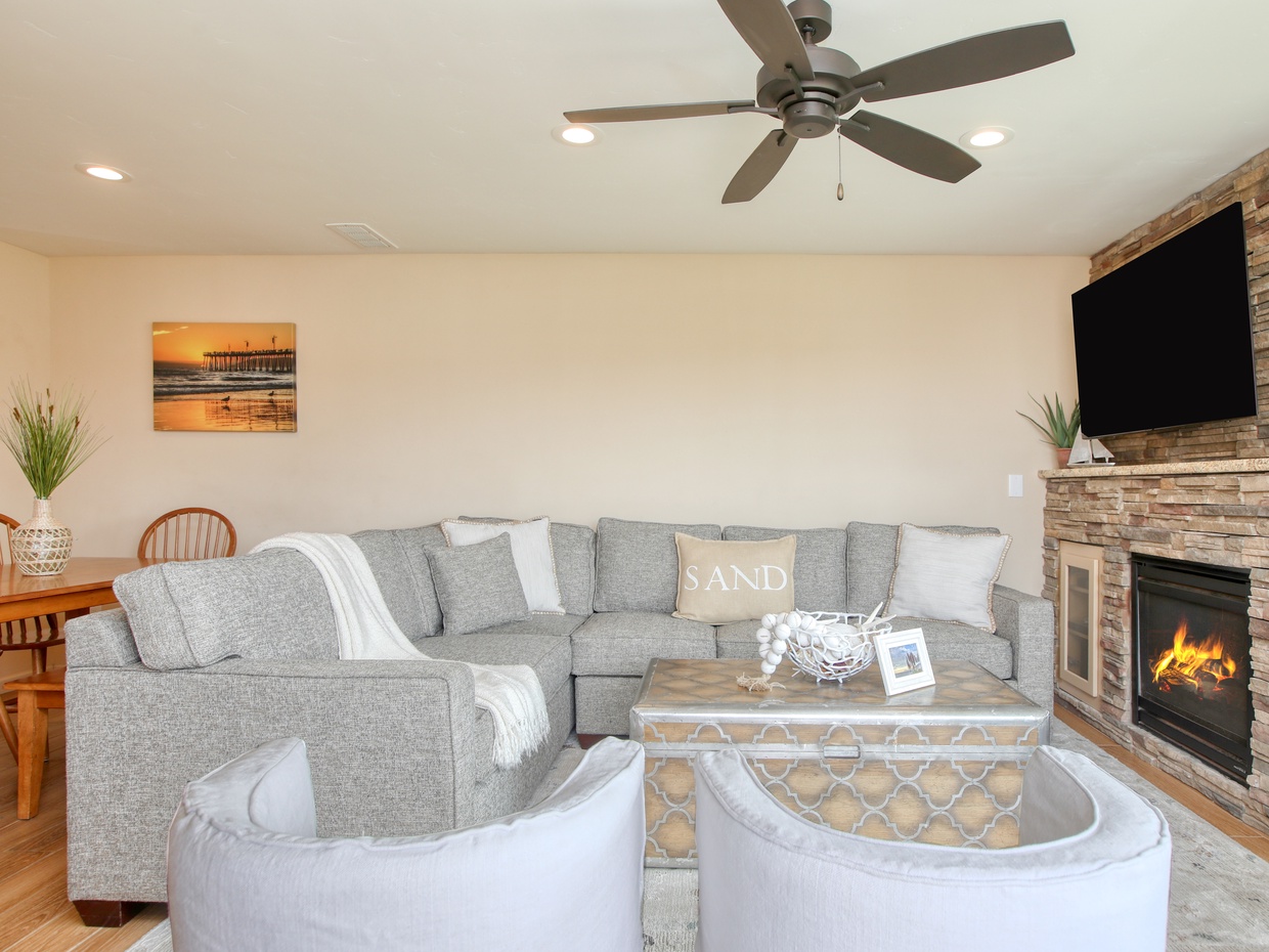The living room is a welcoming space for family and friends, featuring a comfortable sectional and a cozy fireplace. It opens to a patio with two Adirondack chairs, perfect for enjoying morning coffee or evening sunsets. 