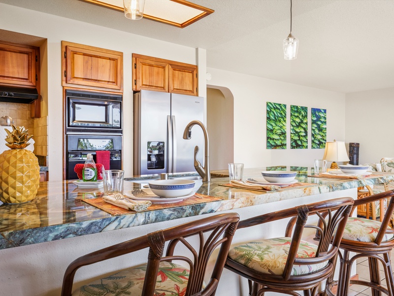 Enjoy a meal or morning coffee at the granite kitchen bar, featuring rattan stools, vibrant decor, and an open view into the living space. A perfect spot to relax and connect