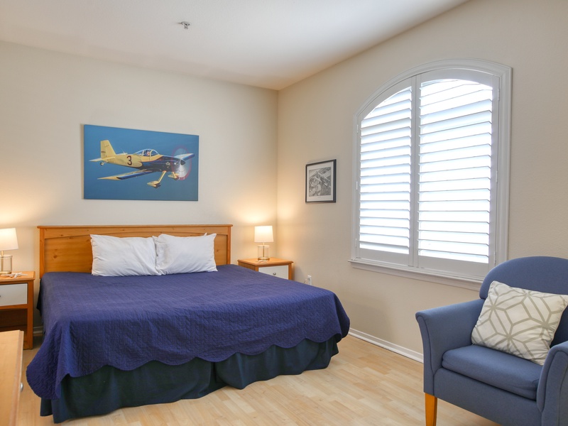 This serene bedroom showcases a plush king bed, soothing blue accents, soft natural light from the arched shuttered window, and a cozy reading chair for relaxing nights.