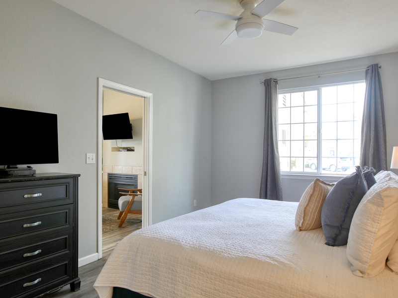 This serene bedroom features a large window that fills the space with natural light, a cozy king sized bed, and a sleek black dresser topped with a flat-screen Smart TV.