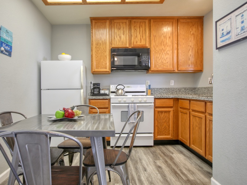 This cozy kitchen features warm wooden cabinetry, a compact dining area with metal chairs, and modern appliances, all set against stylish gray-toned flooring.